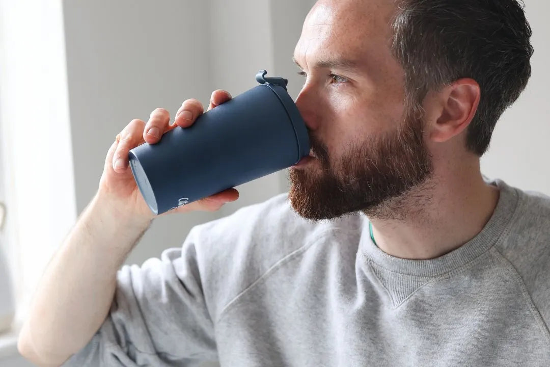 {"image_loaded": true, "load_issue": null, "description": "The image shows a close-up side view of a bearded man wearing a light gray sweatshirt, drinking from a matte navy blue reusable cup with a flip-top lid. The man appears indoors with a neutral-colored background and soft natural lighting coming from a window on the left side. The camera angle is eye-level and focused tightly on the man's face and the cup, creating an intimate feel.", "people": {"count": 1, "roles": ["user"], "visible_demographics": "