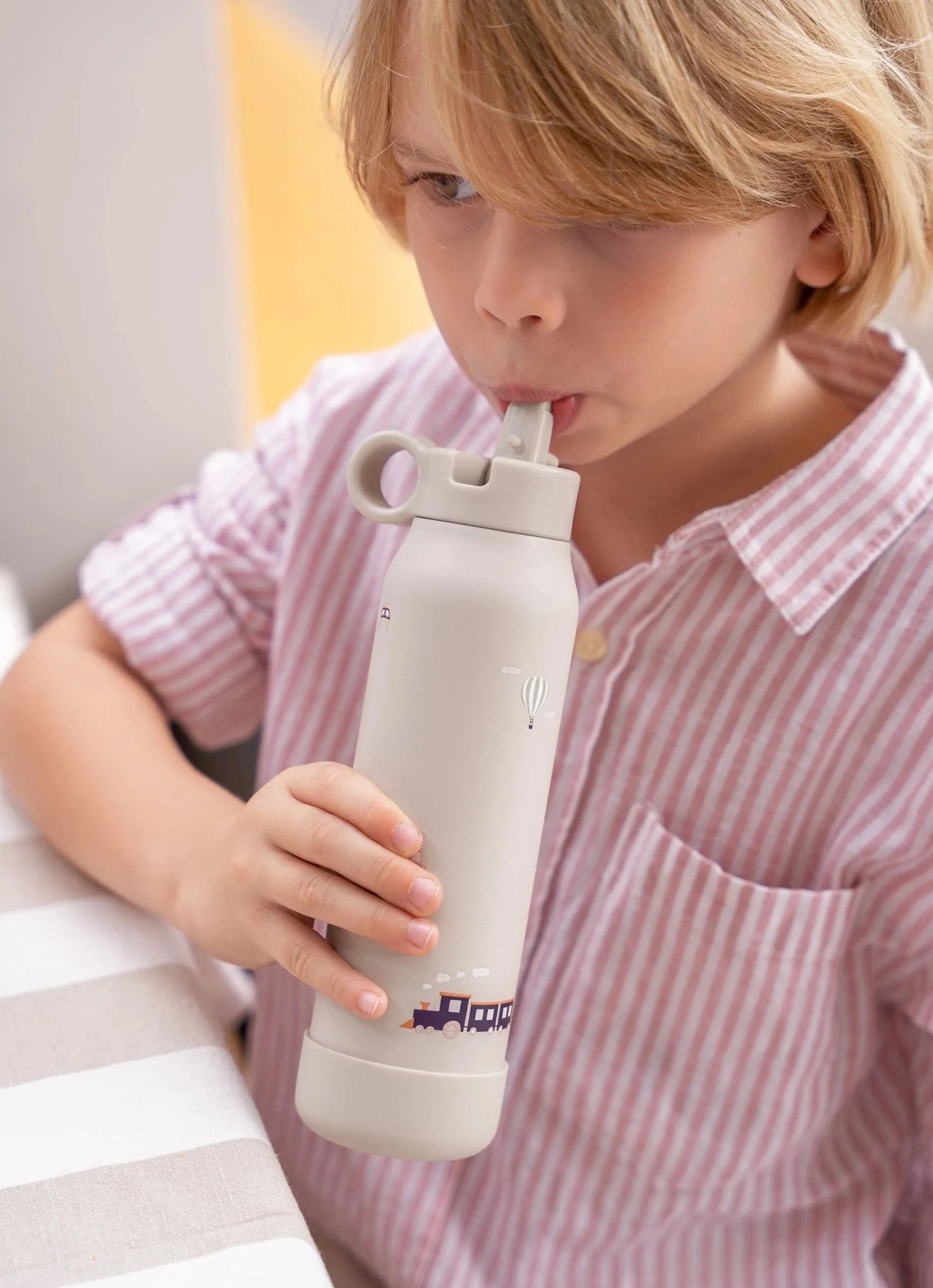 {"image_loaded": true, "load_issue": null, "people": {"count": 1, "roles": ["child"], "visible_demographics": "approximately 4-7 years old, male-presenting, light skin, blonde hair", "attire": "pink and white striped buttoned shirt", "pose_or_activity": "holding and drinking from a bottle with a straw"}, "setting": {"environment_type": "indoor", "location_hints": "table with beige and white striped tablecloth, neutral walls", "depth_scale": "medium", "lighting": "soft, natural or diffused artificial light",